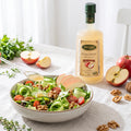 A bottle of Havrano Elma Sirkesi -  apple vinegar stands behind a bowl of fresh green salad with sliced apples, walnuts, and tomatoes on a light tablecloth.